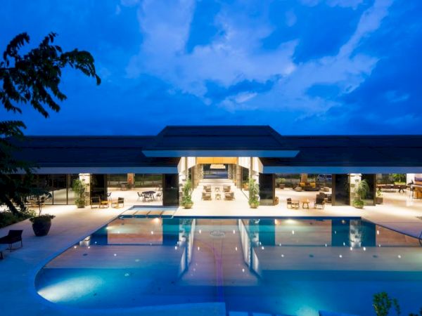 A luxurious modern building with open-air design, featuring a large, illuminated swimming pool, surrounded by lounge chairs, at twilight.