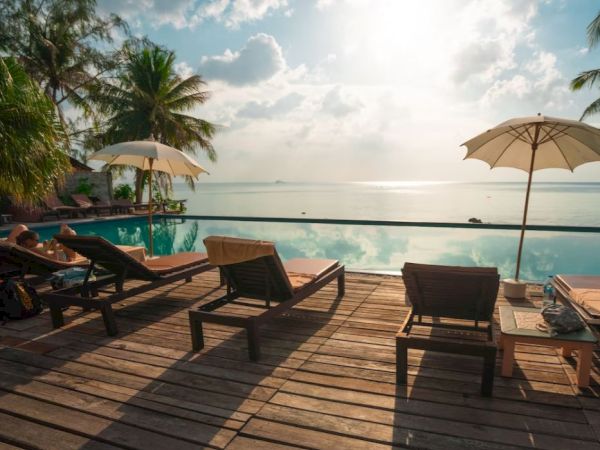 A serene poolside view with lounge chairs, umbrellas, and a wooden deck overlooking a calm sea, surrounded by palm trees, under a clear sky.