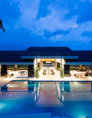 A luxurious, illuminated building with an expansive swimming pool in the foreground under a twilight sky, featuring outdoor seating areas.
