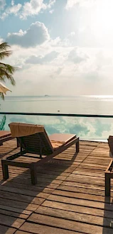 Deck chairs and umbrellas beside an infinity pool overlooking a serene ocean view with palm trees and a partly cloudy sky.