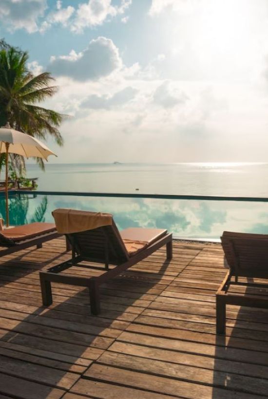 A tranquil poolside scene with lounge chairs and umbrellas overlooking a calm, reflective body of water, surrounded by tropical palm trees.
