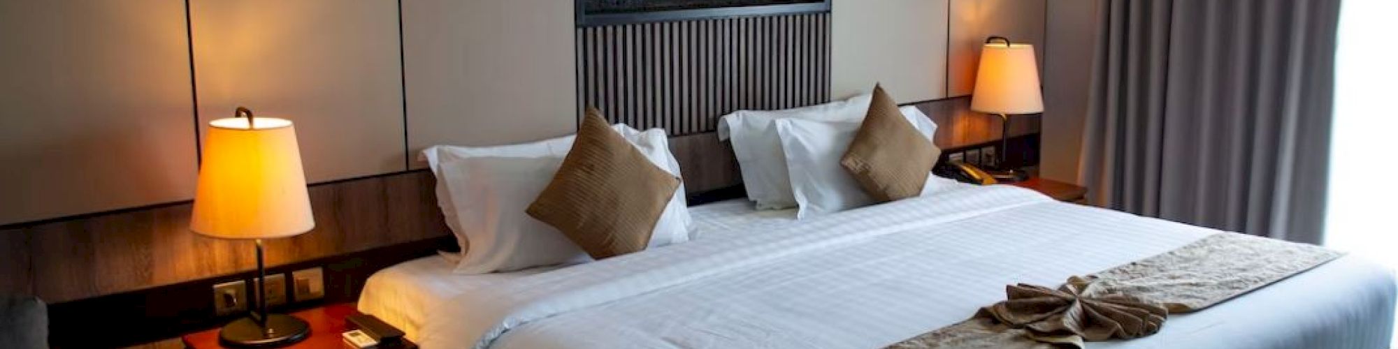 This image shows a neatly designed hotel room with a large bed, two bedside tables with lamps, a small round table, and a wall-mounted decor piece.