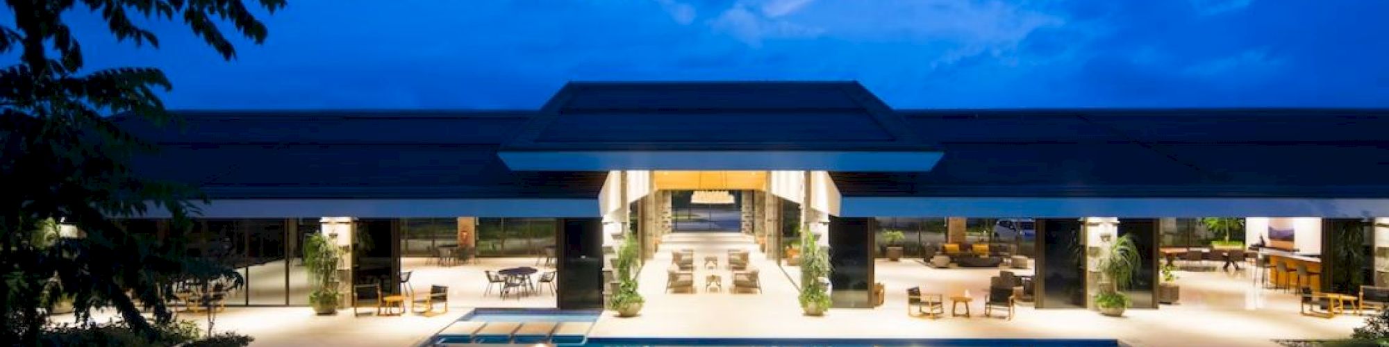 An illuminated modern building with open spaces and seating surrounds a large outdoor swimming pool under a twilight sky.