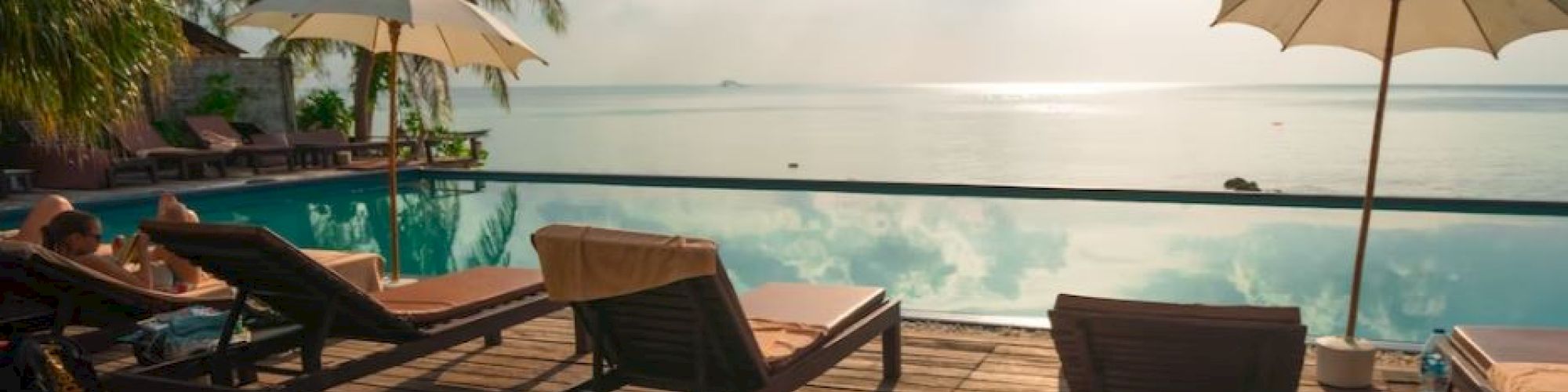 A serene poolside scene with lounge chairs, umbrellas, and a breathtaking view of the sea with the sun shining in a partly cloudy sky.