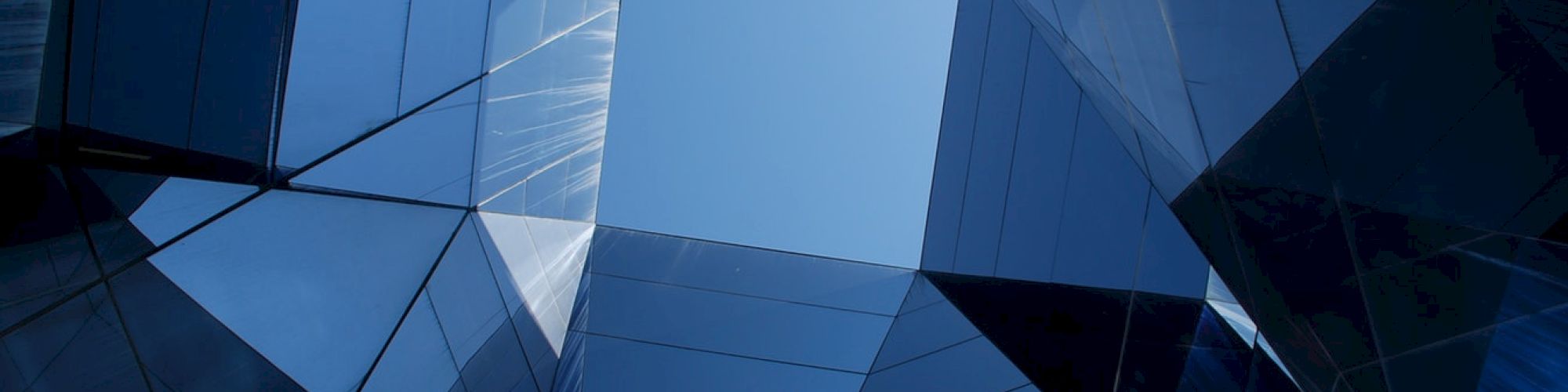 A view looking up through a geometric structure composed of reflective glass panels, showing the sky framed by modern architectural lines.