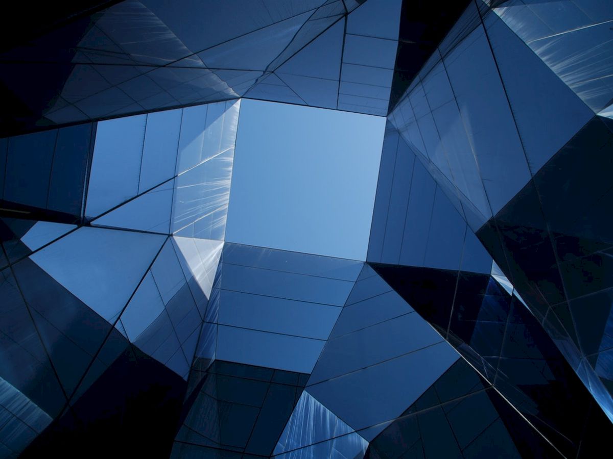 The image shows a view looking up at a geometrically complex architectural structure with blue, reflective glass surfaces and a clear sky above.
