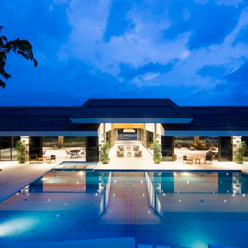The image shows a luxurious resort with a large illuminated swimming pool in the foreground and an open-air building in the background.