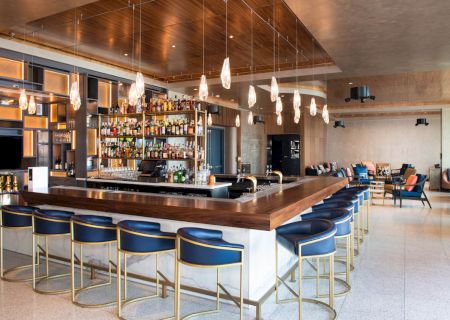 A modern bar with blue and gold stools, a stocked liquor shelf, hanging lights, and a seating area with couches in the background.