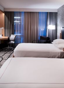A modern hotel room features two neatly made beds, a desk with a chair, a flat-screen TV, and a large window with curtains, ending the sentence.