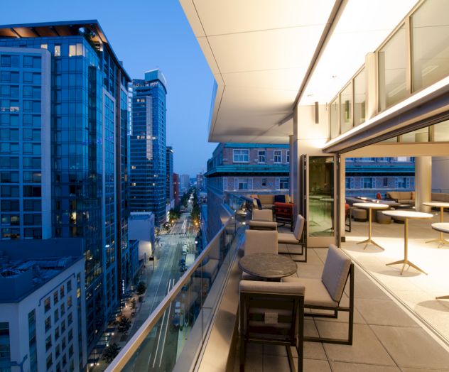 A modern cityscape at twilight, viewed from an upscale balcony with seating and tables, overlooking a vibrant street and tall buildings.