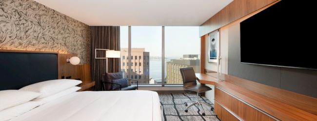 A modern hotel room with a large bed, a desk and chair, a flat-screen TV, and a window with a city view.