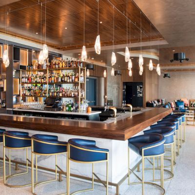 This image shows a modern bar with a long wooden counter, blue bar stools, hanging lights, and a well-stocked bar shelf in a stylish interior space.