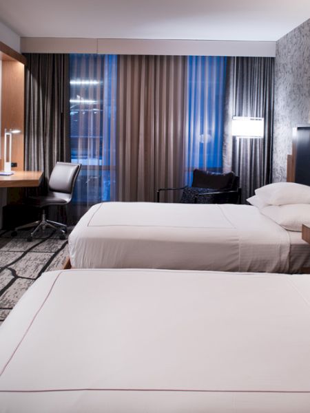 A modern hotel room with two beds, a TV, desk, and window with curtains.