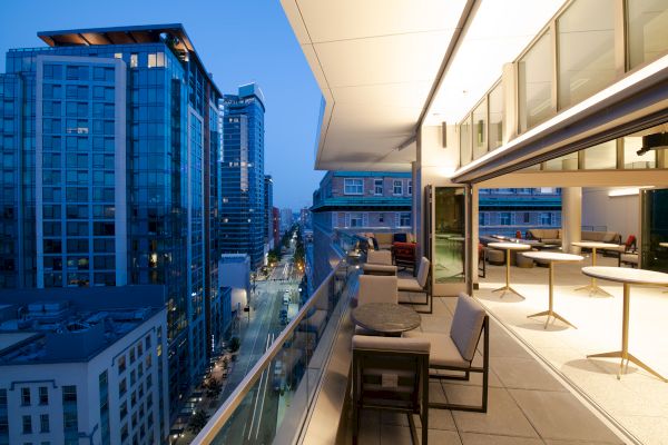 This image shows a modern cityscape with tall buildings and a spacious, furnished balcony overlooking a street at dusk.