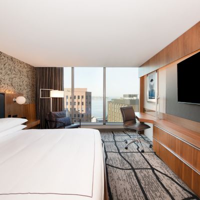 This image shows a modern hotel room with a large bed, a desk and chair, a big flat-screen TV, and a view of buildings outside through large windows.