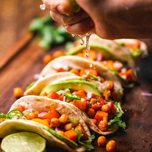 A hand squeezing lime juice over a row of fresh vegetable tacos on a wooden surface, with vibrant ingredients like chickpeas, red onions, and avocado.