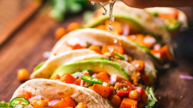 A hand squeezing lime juice over a row of fresh vegetable tacos on a wooden surface, with vibrant ingredients like chickpeas, red onions, and avocado.