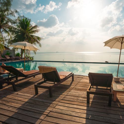 Poolside view with lounge chairs, umbrellas, and palm trees facing a calm ocean under a partly cloudy sky. The wooden deck adds to the relaxing ambiance.