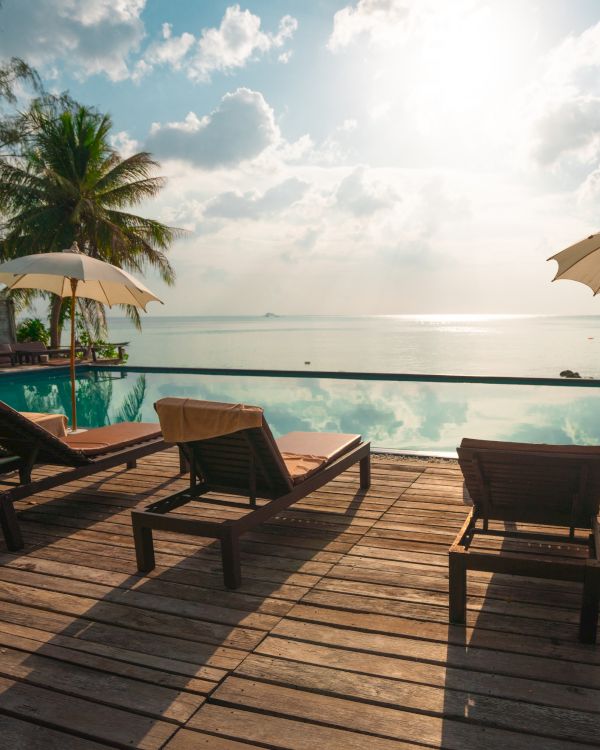 Poolside view with lounge chairs, umbrellas, and palm trees facing a calm ocean under a partly cloudy sky. The wooden deck adds to the relaxing ambiance.