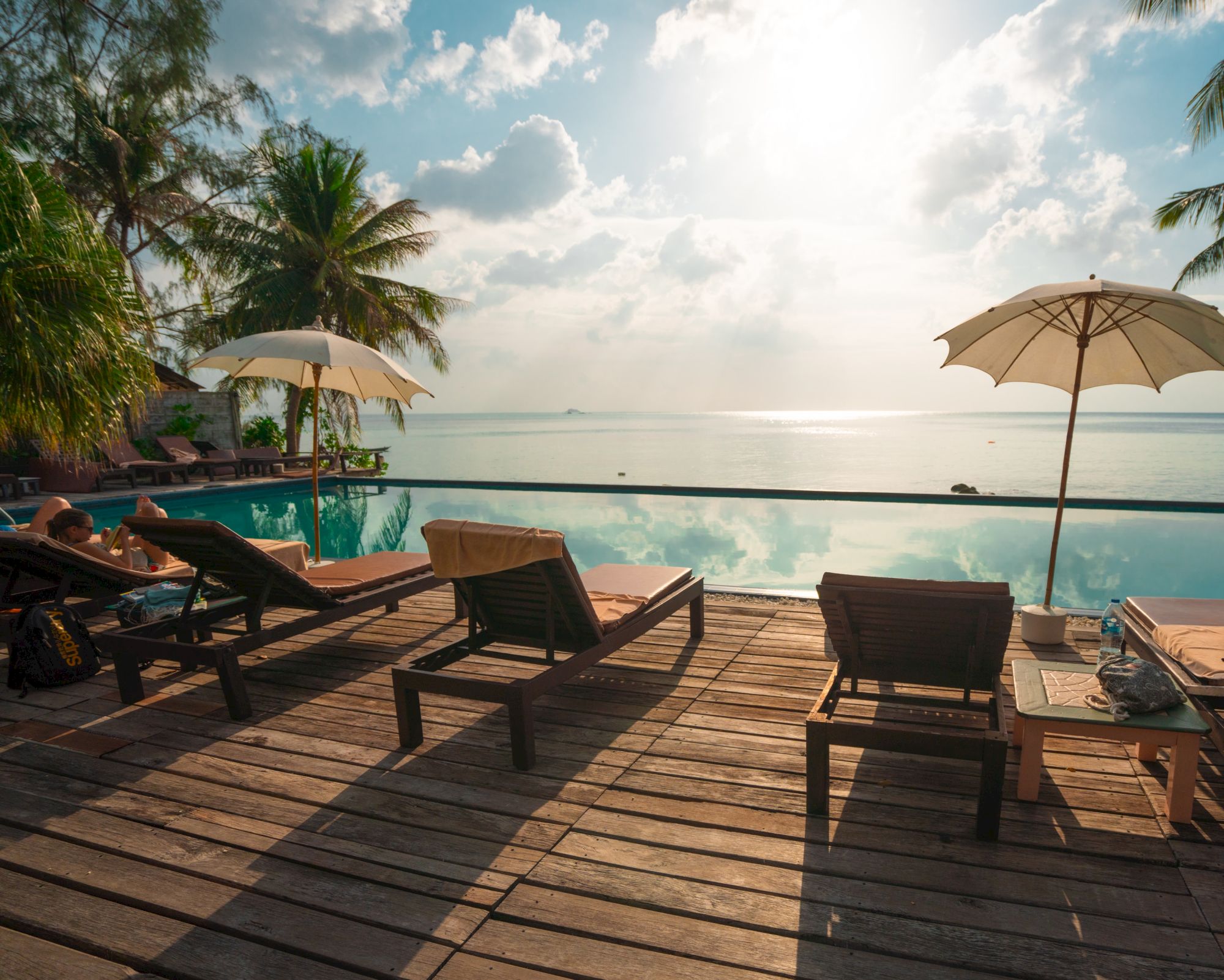 Lounge chairs and umbrellas by a pool are set against a backdrop of a serene ocean view, with palm trees and a beautiful sky completing the scene.