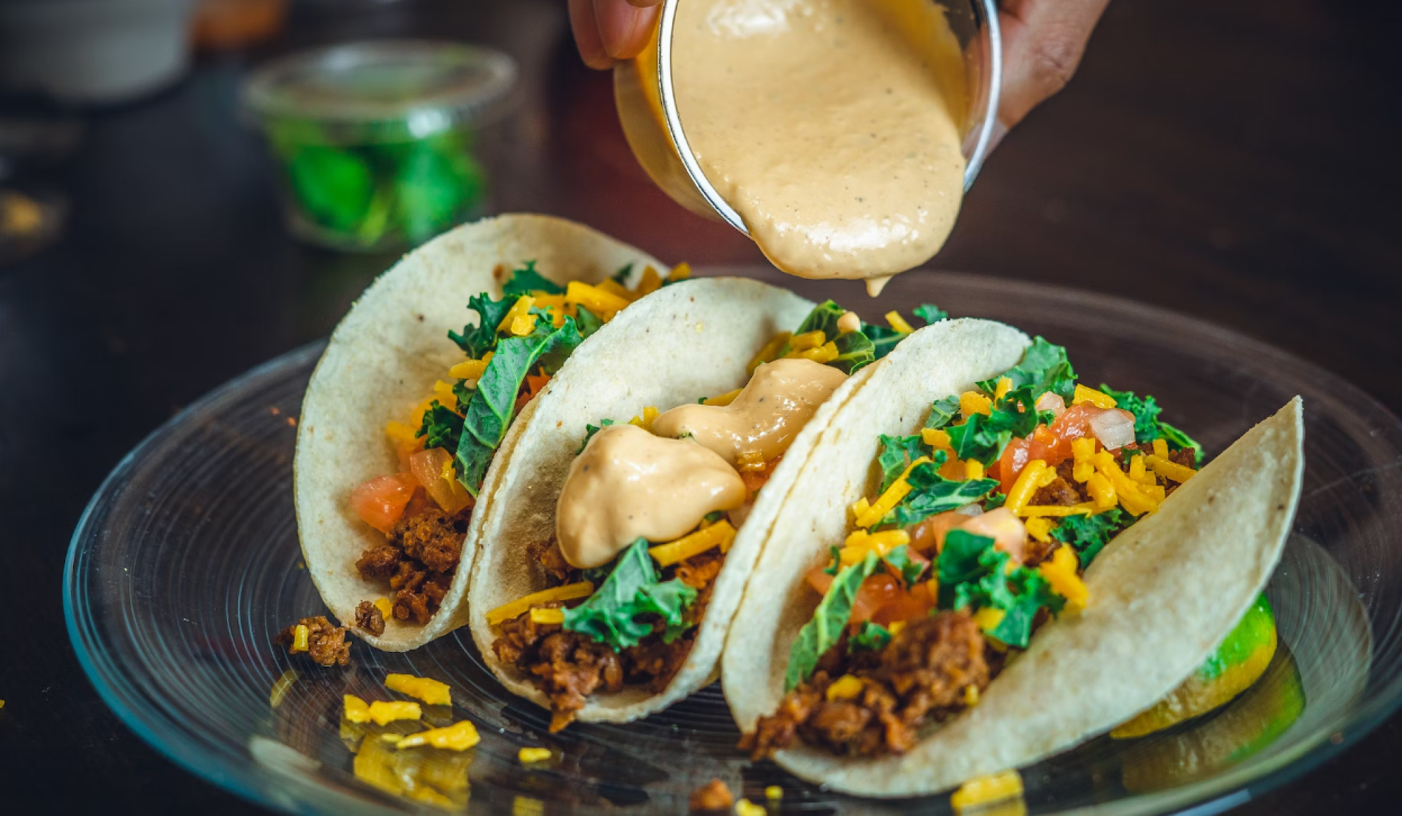 Three tacos on a plate are being topped with a creamy sauce, containing ingredients like ground meat, lettuce, tomato, and shredded cheese.