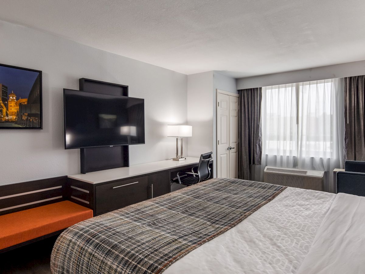 A modern hotel room featuring a bed, flat-screen TV, desk, chair, sofa, and window with curtains. The room is well-lit and has a cozy ambiance.