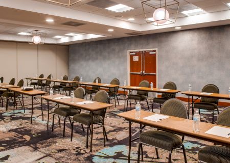 A conference room with rows of tables and chairs, each table set with notebooks and water bottles, under a ceiling with hanging lights.