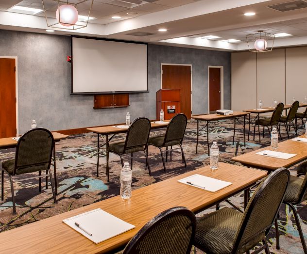 A conference room with tables and chairs arranged classroom-style, facing a screen and podium. Water bottles and notepads are placed on the tables.