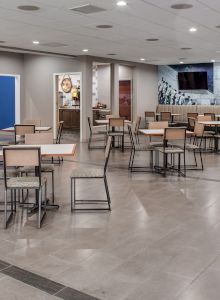 The image shows a modern, clean cafeteria with empty tables and chairs, a counter with high stools, and a mural on the back wall.