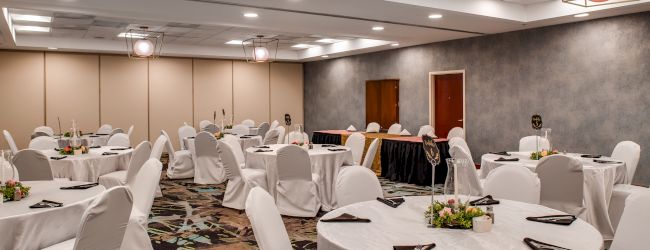 A decorated banquet hall with round tables covered in white tablecloths and chairs, floral centerpieces, and black napkins on each table, ends the sentence.