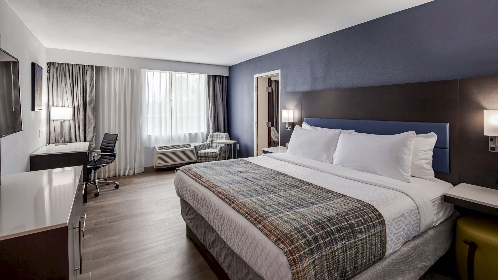 This image shows a modern hotel room with a king-size bed, desk, chair, and large window with curtains. The room has a clean, minimalist design.