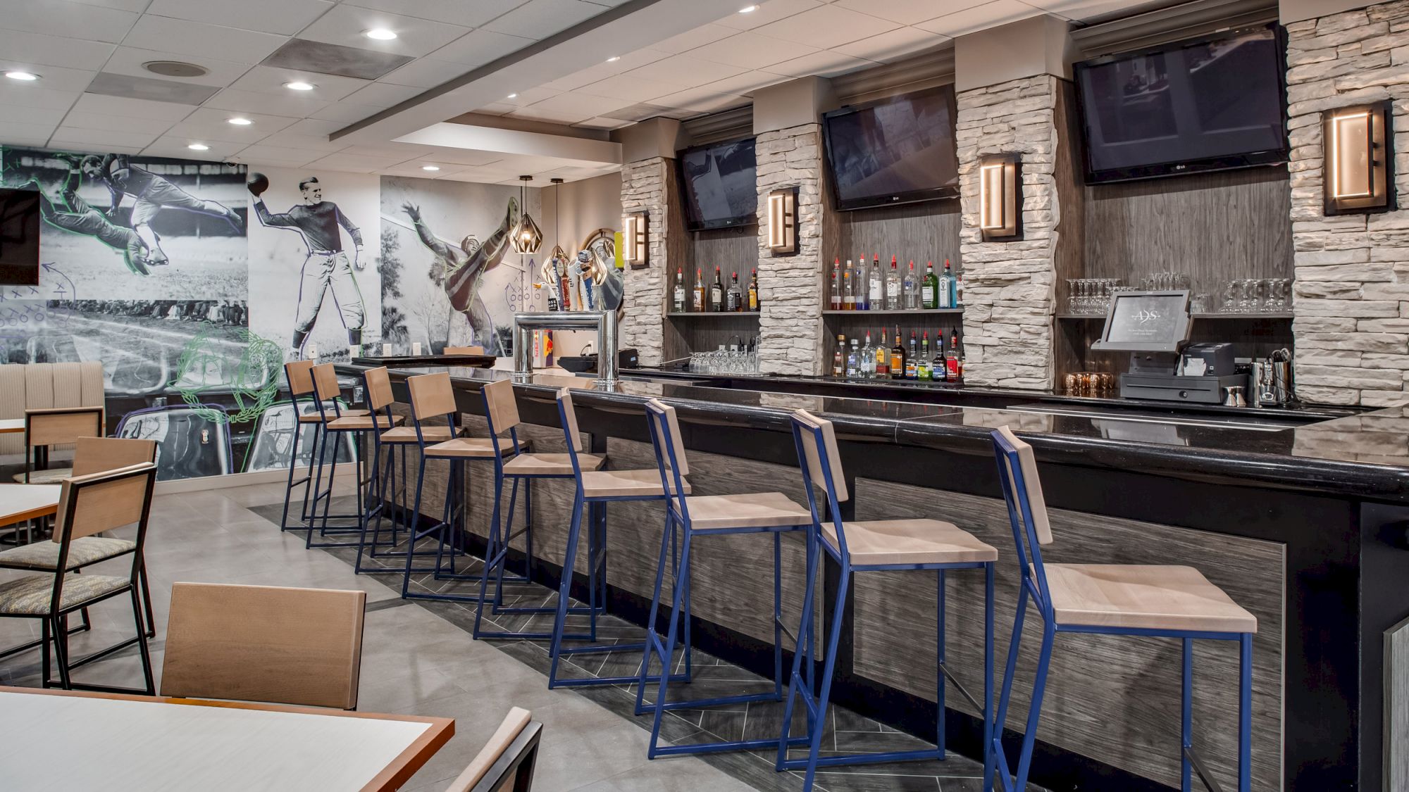 A modern bar area with high chairs, a well-stocked drink shelf, multiple flat-screen TVs, and sports-themed wall art.