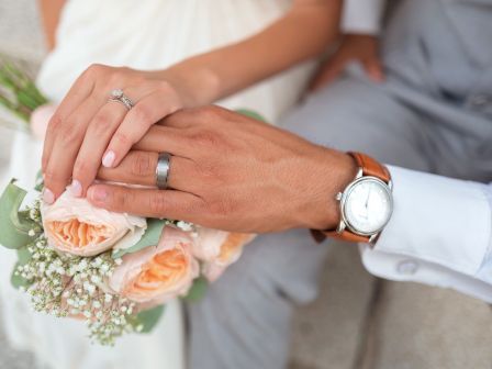 A couple's hands are shown, each wearing wedding rings, with the woman holding a bouquet of roses and baby's breath.