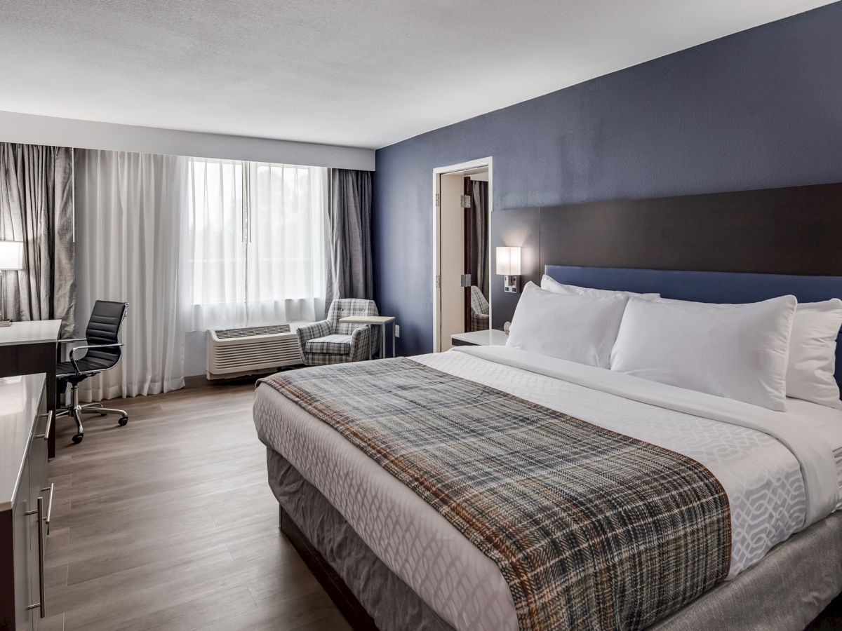 A modern hotel room with a large bed, plaid blanket, TV, desk, office chair, armchair by the window, air conditioner, and wall lights.