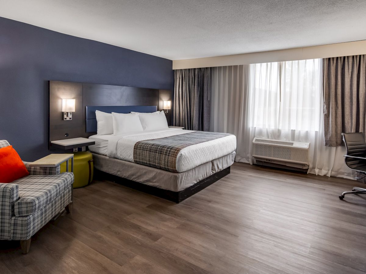 This image shows a modern hotel room with a bed, desk, armchair, lamps, and a window with curtains. The decor is simple and clean.