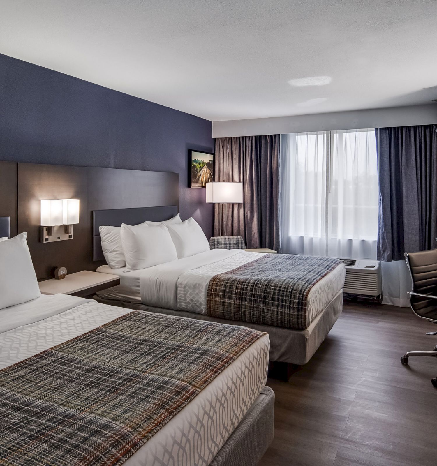 The image shows a hotel room with two double beds, a desk with a chair, a TV, and a window with curtains letting in natural light.