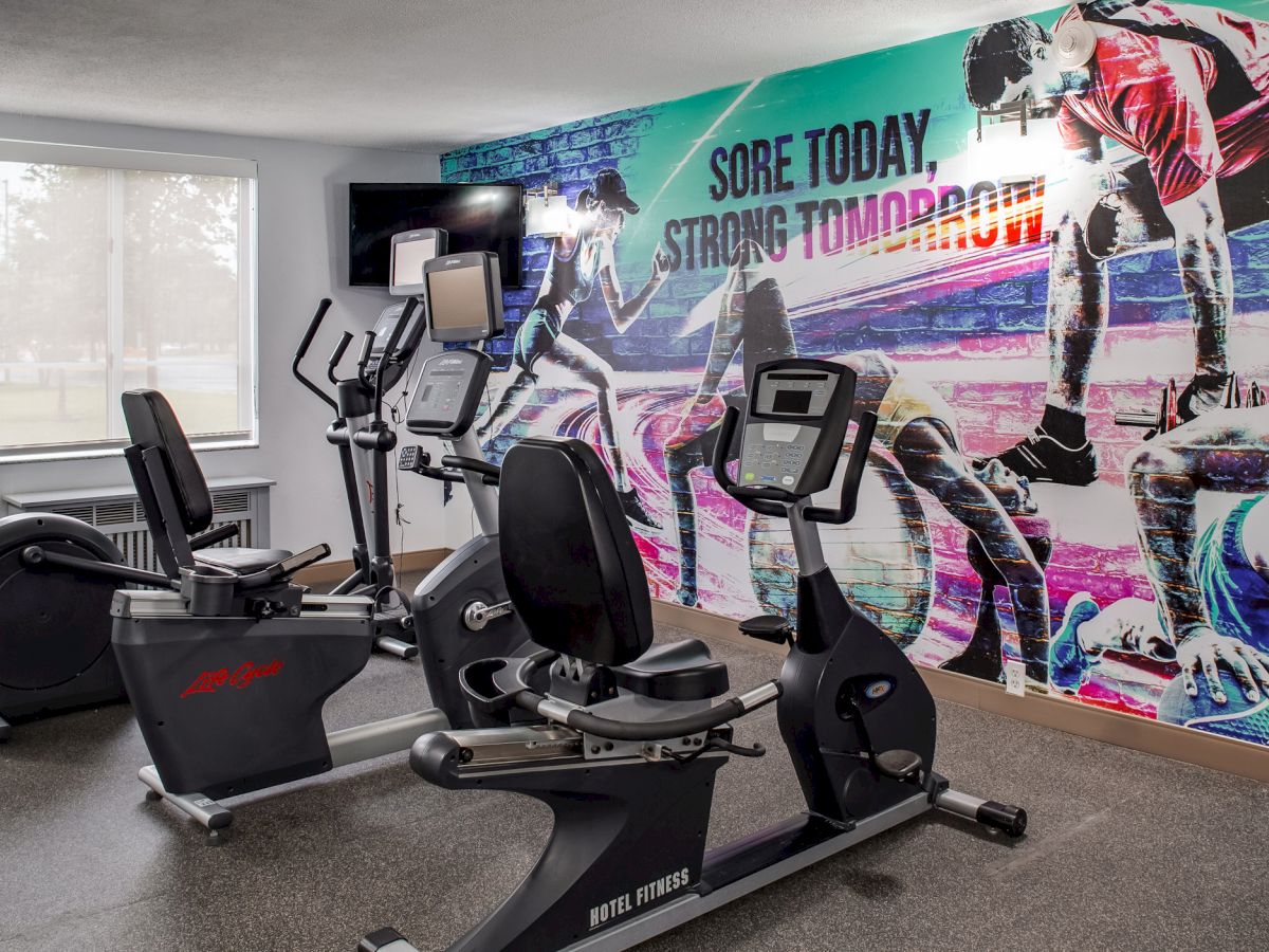 A small gym with two stationary bikes and an elliptical machine. There's a colorful mural with athletes and the text 