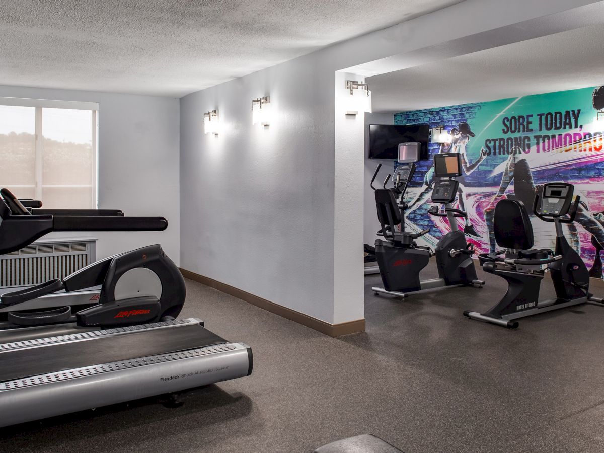 The image shows a gym with treadmills, exercise bikes, and a motivational mural that reads "Sore today, strong tomorrow."