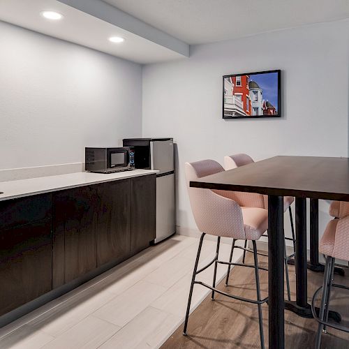 The image depicts a small kitchenette area with a sink, microwave, and mini fridge, adjacent to a high table with four upholstered chairs.