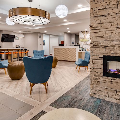 The image shows a modern lobby area with blue chairs, a reception desk, a stone fireplace, and a bar area in the background.