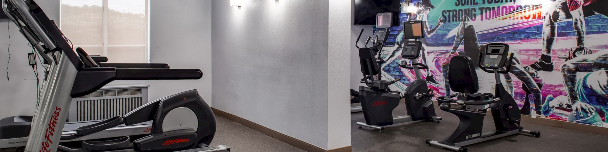 The image shows a gym with treadmills, exercise bikes, and a motivational mural on the wall saying, "Sore today, strong tomorrow."