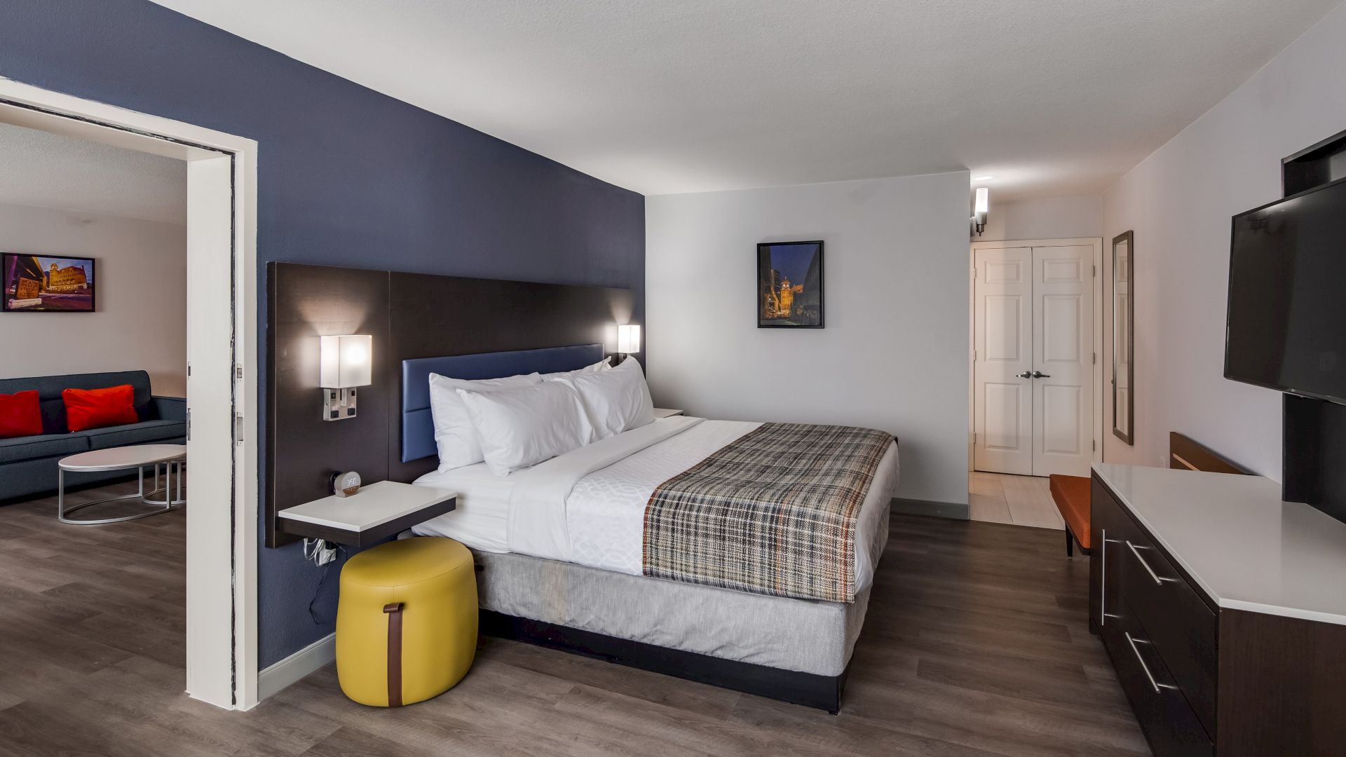 A modern hotel room with a bed, wall art, flat-screen TV, dresser, and adjacent living area with a couch and red pillows.