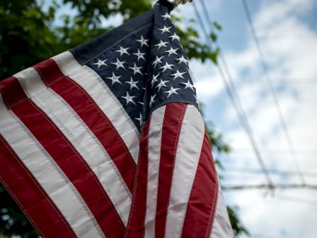 An American flag is flying against a background of trees, blue sky, and power lines.