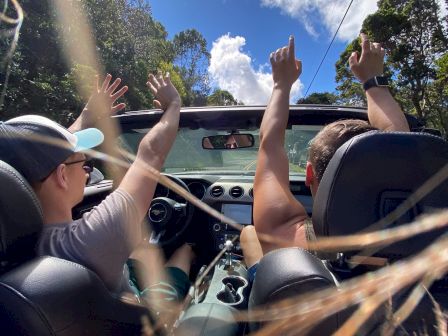 Two people in a convertible car with the top down are raising their hands, driving on a sunny day with blue skies and trees around.