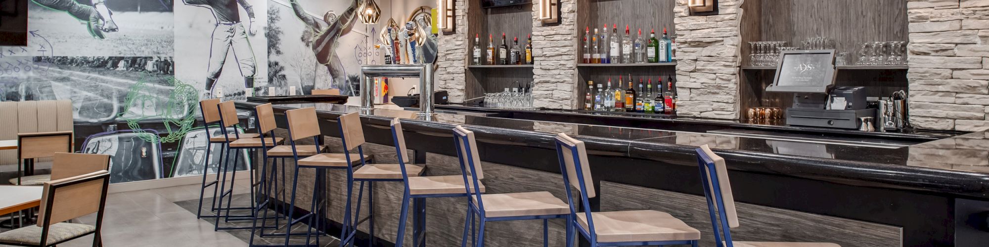 The image shows a modern bar with high chairs, a counter, and a variety of drinks displayed on shelves. There are sports images on the walls.