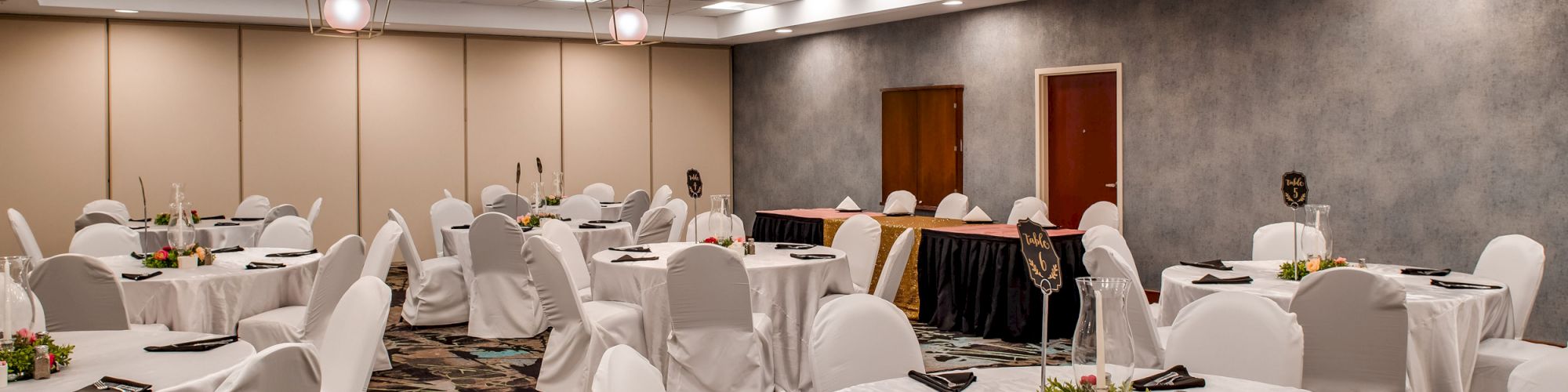 A banquet hall is set up with round tables covered in white cloth, decorated with centerpieces, and black napkins, ready for an event.