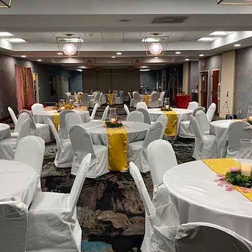 The image shows a banquet hall set up with round tables covered in white cloths, chairs with white covers, and yellow table runners.