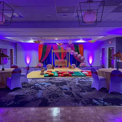 A decorated banquet hall with purple lighting, round tables with chairs, and a colorful setup in the center, likely for an event or celebration.