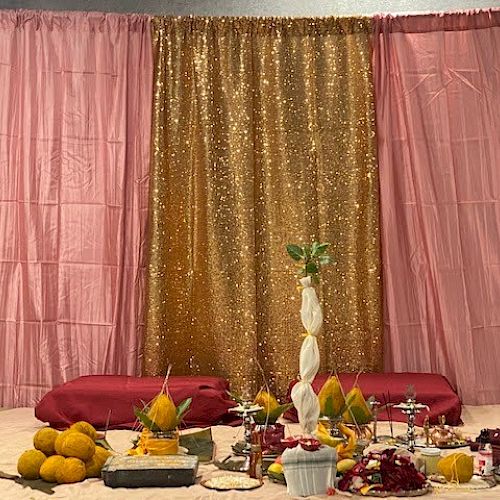 The image shows a festive setup with a pink and gold backdrop, various ceremonial items, fruits, and flowers arranged in the foreground.