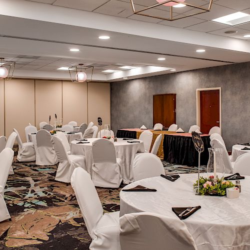 A banquet hall set up with round tables and white chairs, adorned with centerpieces and neatly placed napkins, ready for an event.